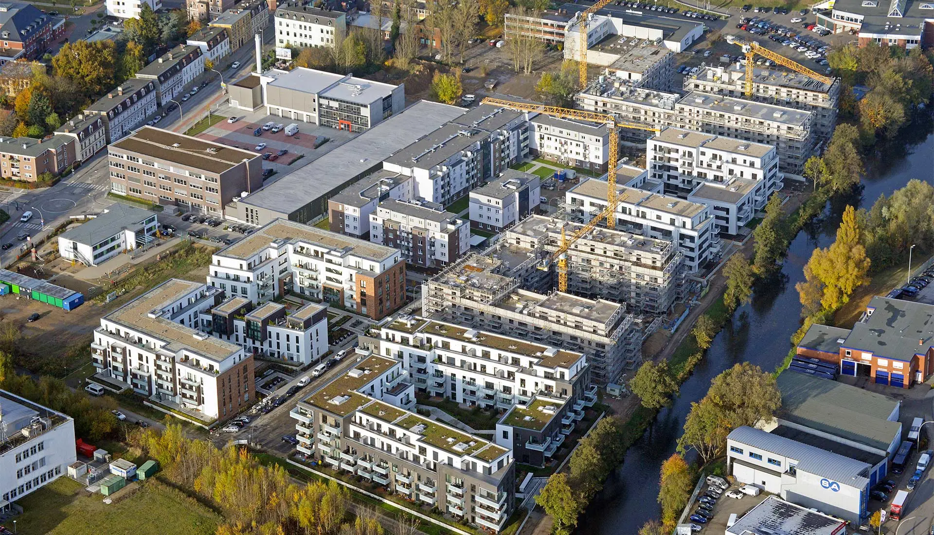 Luftaufnahme einer städtischen Siedlung mit vielen weißen Wohngebäuden, einigen im Bau mit Kränen, und einem Fluss.