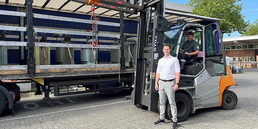 Ein Mann steht neben einem Gabelstapler, während ein Fahrer im Fahrzeug sitzt, im Hintergrund sind Paletten und einen Transporter sichtbar.