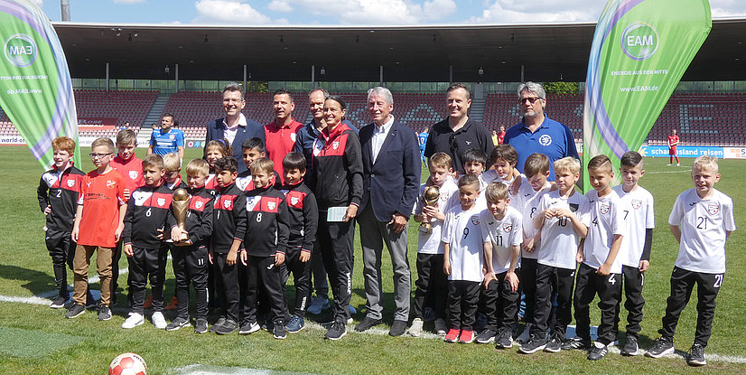 Eine Gruppe von Erwachsenen und Kindern in Sportkleidung posiert auf einem Fußballfeld, umgeben von zwei großen Bannern.