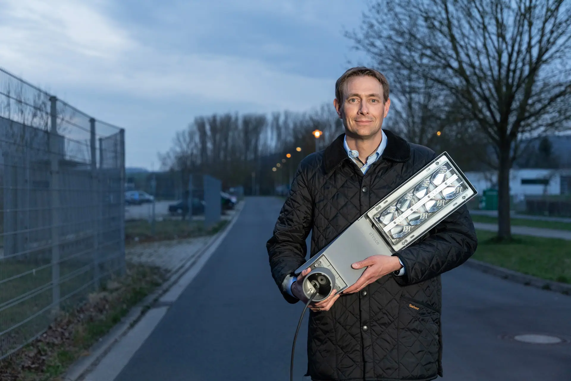  Ein Mann in dunkler Jacke hält eine moderne LED-Straßenlaterne in den Händen, im Hintergrund eine Straße bei Dämmerung.