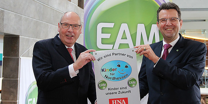 Zwei Männer in Anzügen halten ein Schild mit der Aufschrift Wir sind Partner von Kinder für Nordhessen"" in einer modernen Umgebung.