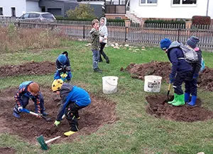 Sechs Kinder buddeln im Garten, während zwei andere im Hintergrund stehen. Erde wird mit Schaufeln bearbeitet.