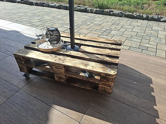 Holzpalette als Tisch im Freien, mit Glas offen, einem Teller und einem Lampenschirm auf einem gepflasterten Boden.
