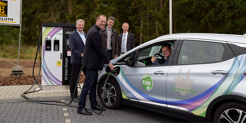 Verschiedene Personen stehen vor einer Schnellladestation für Elektrofahrzeuge, während eines Ladeprozesses.