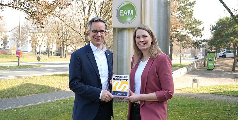 Zwei Personen stehen im Freien vor einem EAM‑Schild und halten gemeinsam eine Auszeichnung mit der Aufschrift „Top 2026 Company – kununu“. Im Hintergrund sind eine Straße, Bäume, ein Gehweg und eine Ladeinfrastruktur für Elektrofahrzeuge zu sehen.