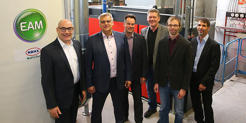 Gruppenfoto von sechs Personen vor einer Maschinenanlage, mit EAM- und NBHX-Logos im Hintergrund.