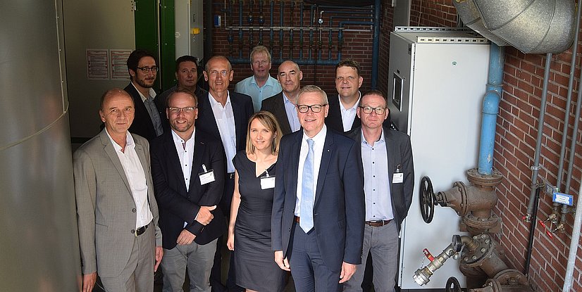 Gruppenfoto von zehn Personen in einem technischen Umfeld, einige tragen Namensschilder. Hintergrund mit Rohren und Anlagen.