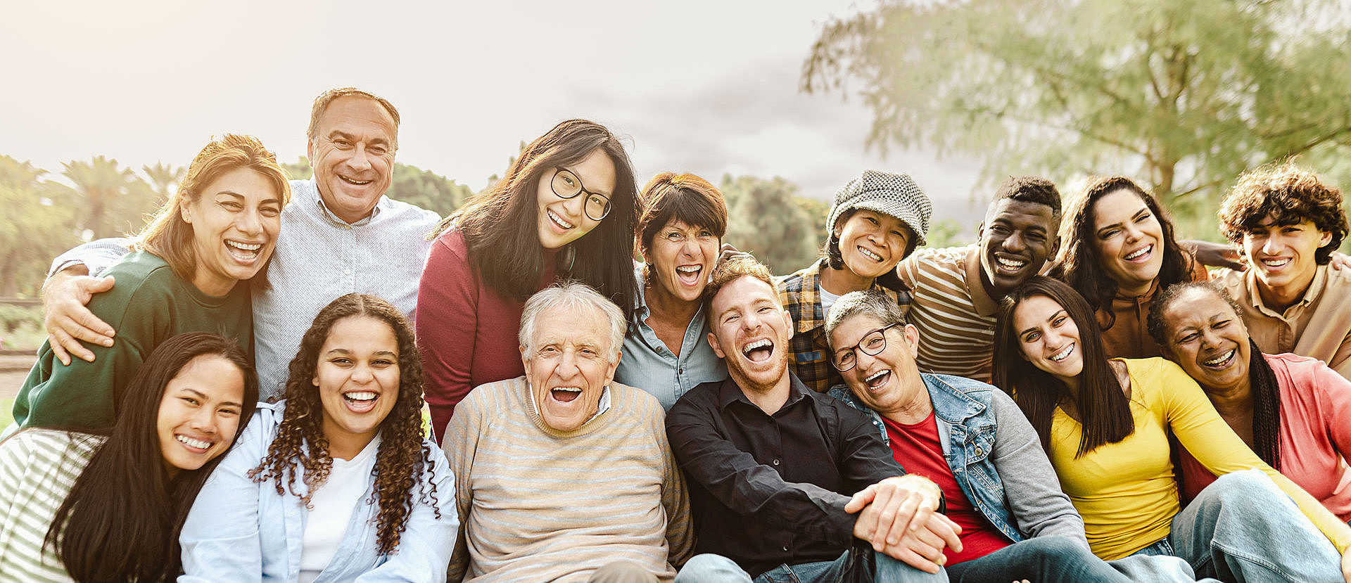 Gruppe diverser Menschen unterschiedlichen Alters, die eng zusammenstehen, lachen und in die Kamera schauen.