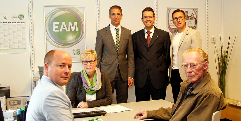 Gruppe von fünf Personen in einem Büro, am Tisch sitzend und stehend, mit einem EAM-Logo im Hintergrund.