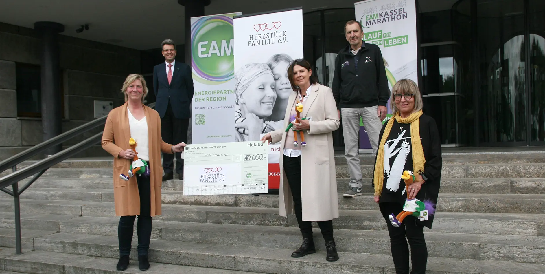 Gruppenfoto von vier Personen, die einen Scheck für eine gemeinnützige Initiative präsentieren, vor einem Gebäude stehend.