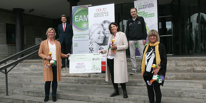 Gruppenfoto von vier Personen, die einen Scheck für eine gemeinnützige Initiative präsentieren, vor einem Gebäude stehend.