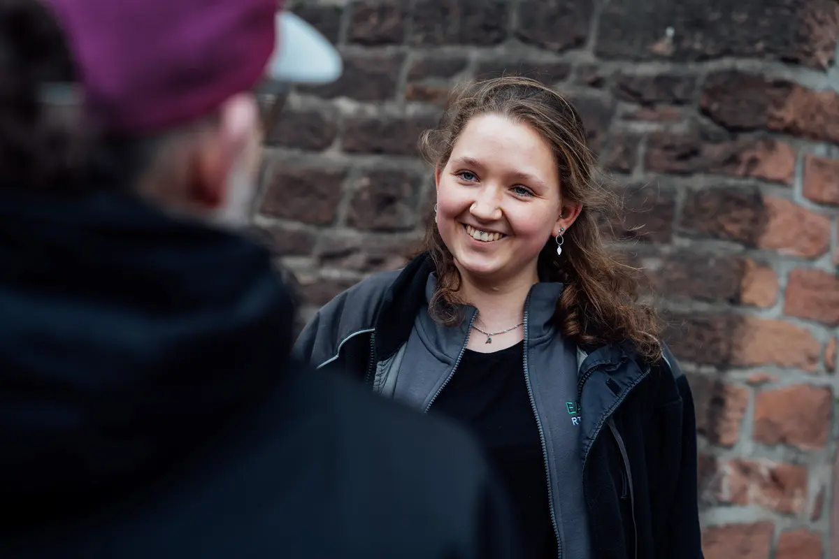 Lächelnde Frau mit lockigem Haar in einem grauen Jackett, im Gespräch vor einer steinernen Wand.