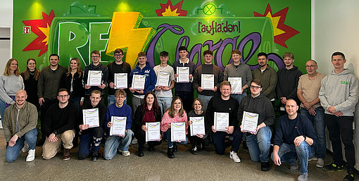 14 junge Nachwuchskollegen stehen mit ihren Zeugnissen in der Hand in einem bunten Raum im EAM Campus, bei ihnen stehen die Leiterin der Aus- und Weiterbildung Miriam Franz, der Leiter der gewerblich-technischen Ausbildung Anthony Speer und Ausbilderin Vanessa Hartung. Auch die weiteren Aus- und Weiterbilder sowie die Pädagoginnen waren bei der Zeugnisübergabe dabei und gratulierten den jungen Kolleginnen und Kollegen herzlich.