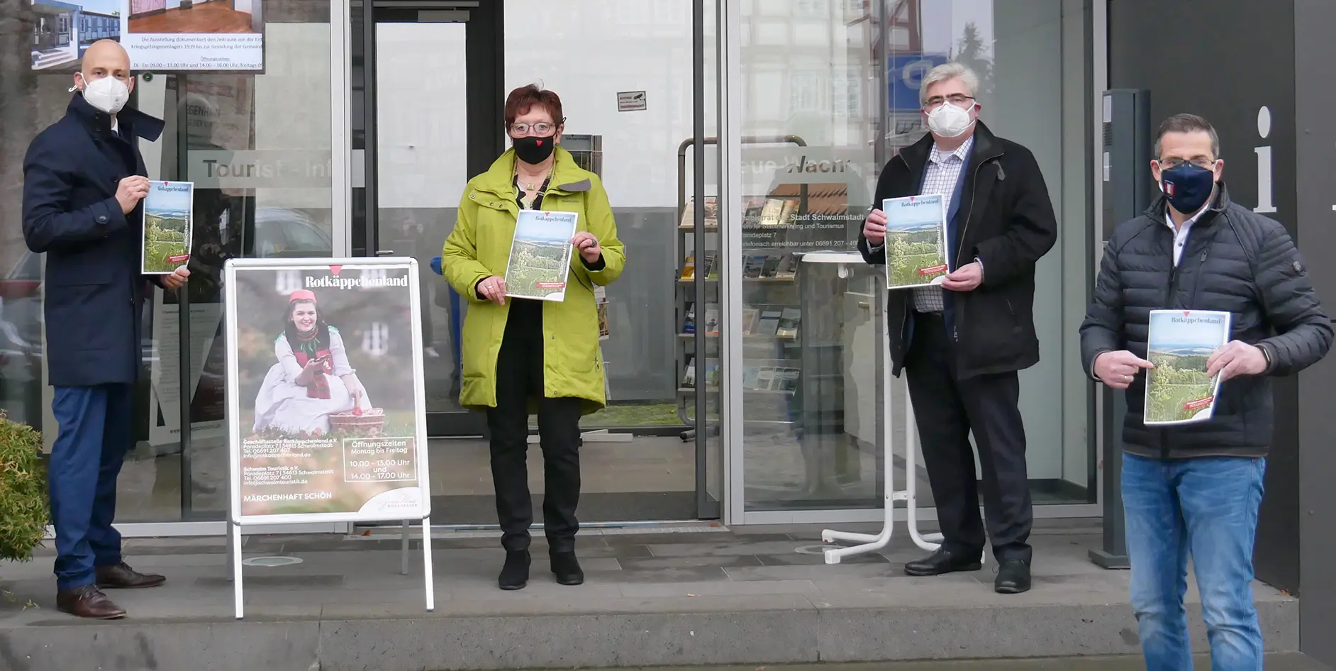 Vier Personen stehen vor einer Touristeninformation, halten Broschüren. Plakat für Rotkäppchenland im Hintergrund.