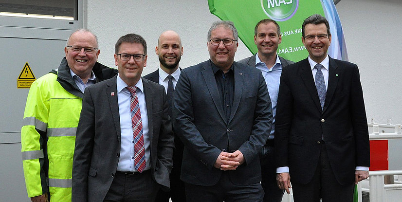Gruppe von sechs Personen im Freien, lächelnd, mit einem Schild im Hintergrund. Sicherheitswarnung an der Wand sichtbar.