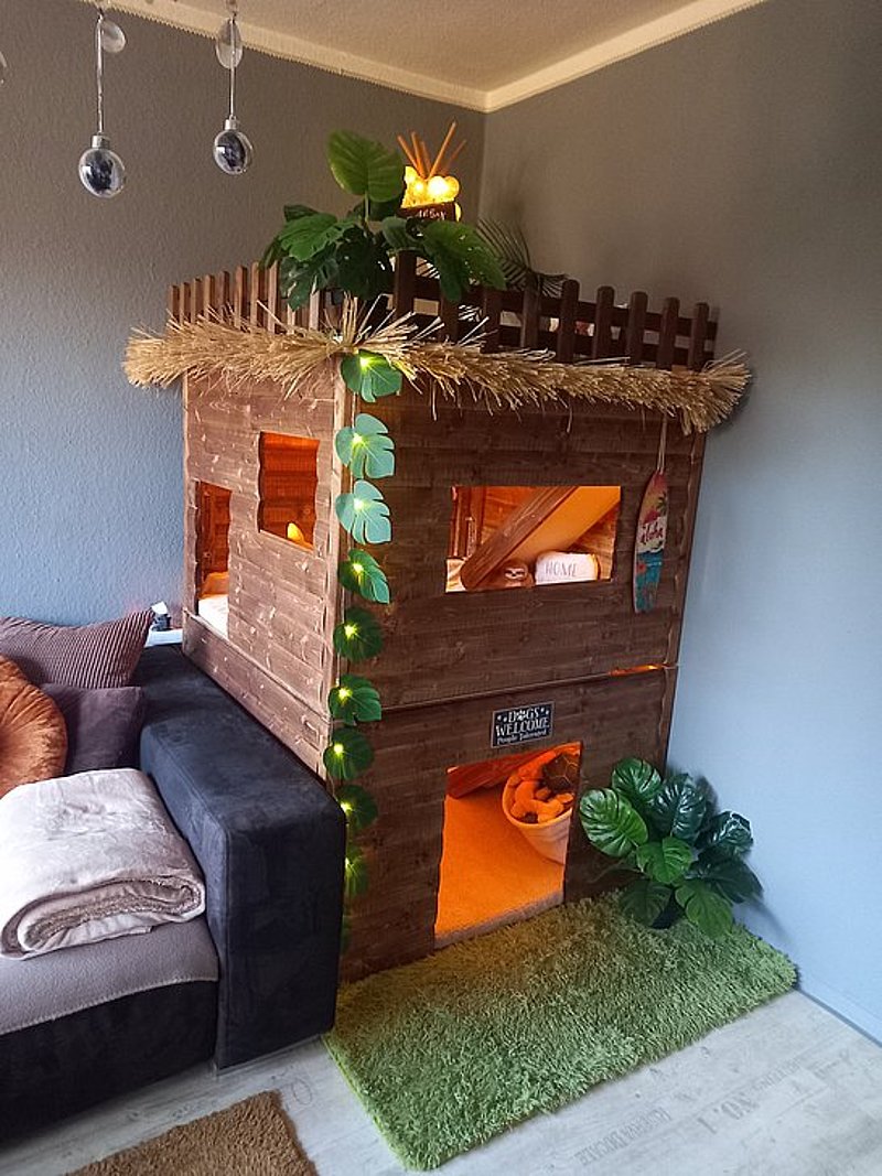 Holzspielhaus mit Dach und Pflanzen, dekoriert mit Lichtern, neben einem Sofa und Teppich in moderner Wohnumgebung.
