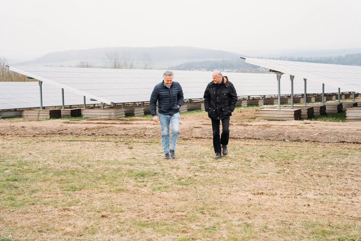 Zwei Personen gehen an Solarpanelen vorbei, umgeben von einer grünen Wiese unter einem grauen Himmel.