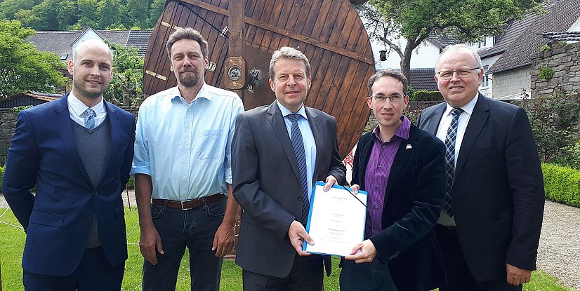 Fünf Männer stehen in einem Garten, lächeln und halten ein Dokument in der Hand. Im Hintergrund eine große Holzstruktur.