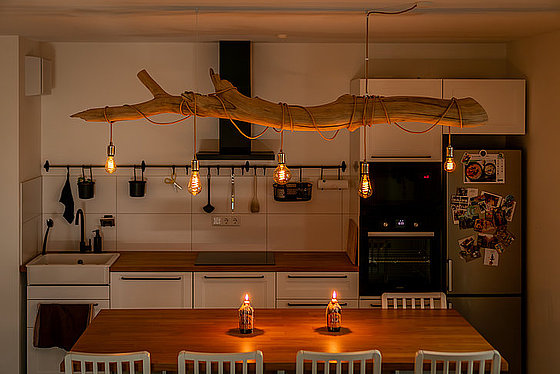 Moderne Küche mit Holzdecke, hängenden Lampen und einem großen Tisch mit Kerzen. Minimalistisches Design und warmes Licht.