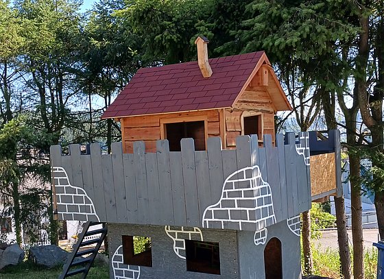 Ein Spielhaus aus Holz mit rotem Dach, umlaufender Veranda und Fenstern, umgeben von Bäumen.