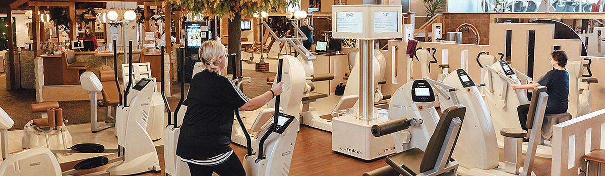 Fitnessstudio mit modernen Trainingsgeräten, Menschen beim Trainieren und einem begrünten Bereich im Hintergrund.