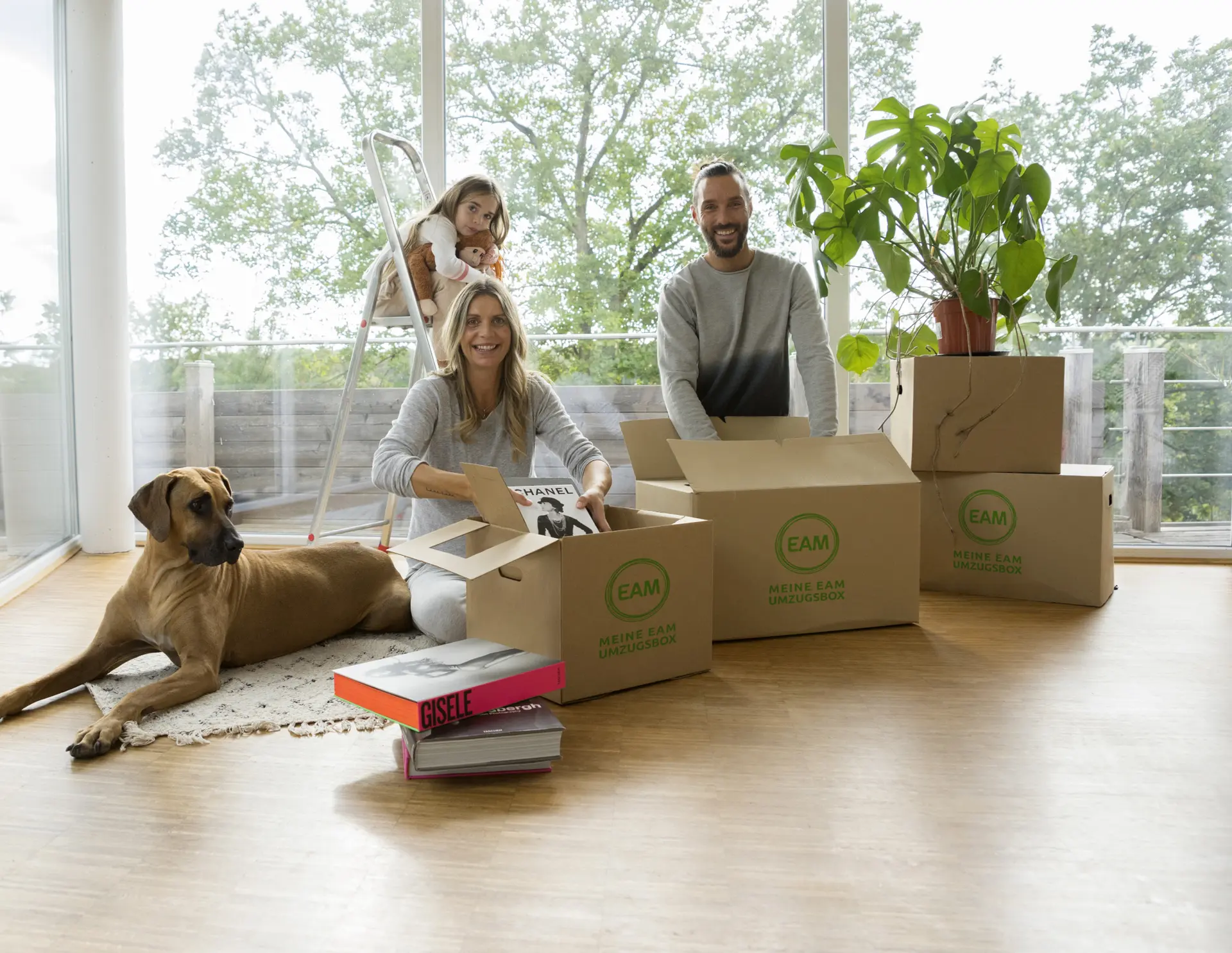  Ein glückliches Paar und ein Kind beim Auspacken von Umzugskartons, wobei ein Hund entspannt auf dem Boden liegt.