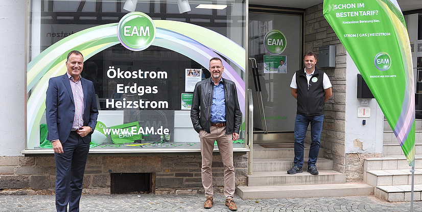 Drei Männer stehen vor dem EAM-Kundenservicebüro in Melsungen, umgeben von Werbematerialien für Ökostrom, Gas und Heizstrom.