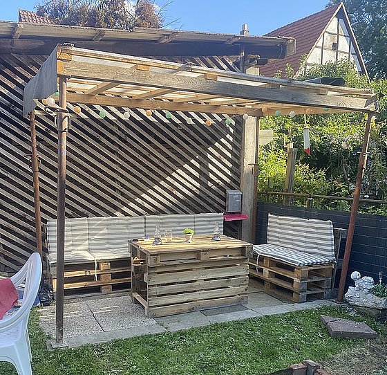 Gemütlicher Gartenbereich mit Holzmöbeln, Sonnenschutz und grüner Umrandung.