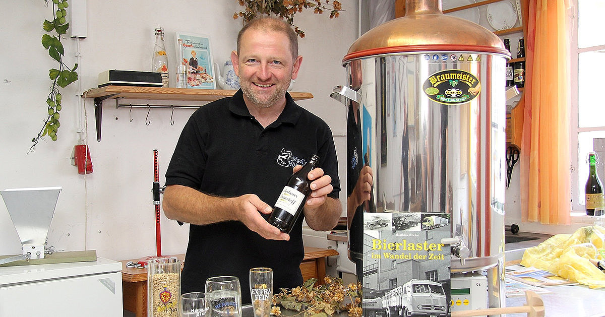 Ein Mann in einer Brauerei hält eine Flasche Bier, umgeben von Gläsern, Zutaten und Brauerei-Materialien.