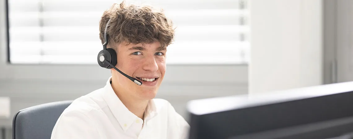 Junger Mann mit Headset, lächelt, sitzt am Schreibtisch vor einem Computerbildschirm, helle Büroumgebung.