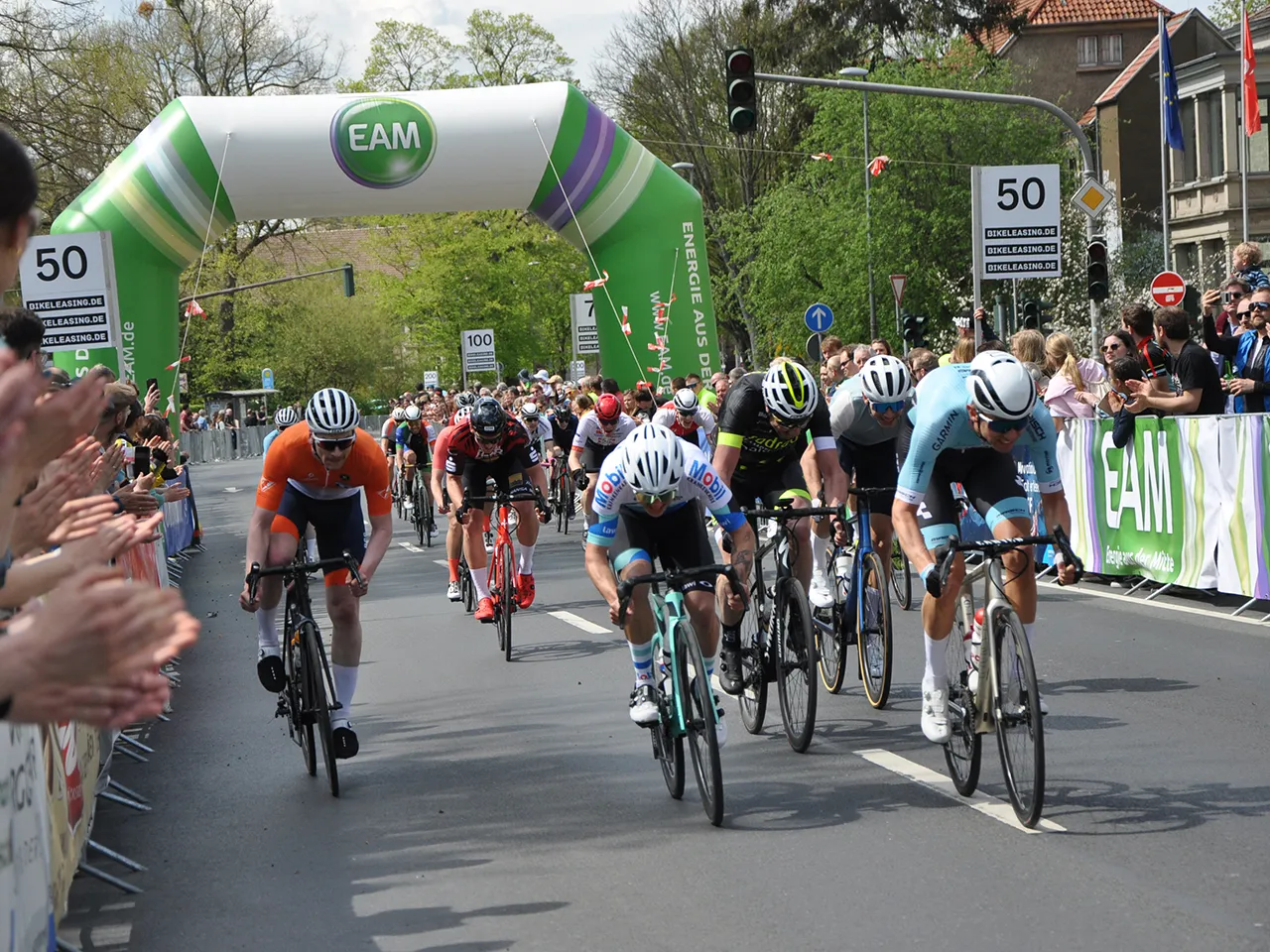 Radrennen mit mehreren Fahrern, die um einen Zielbogen fahren, während die Zuschauer enthusiastisch applaudieren.
