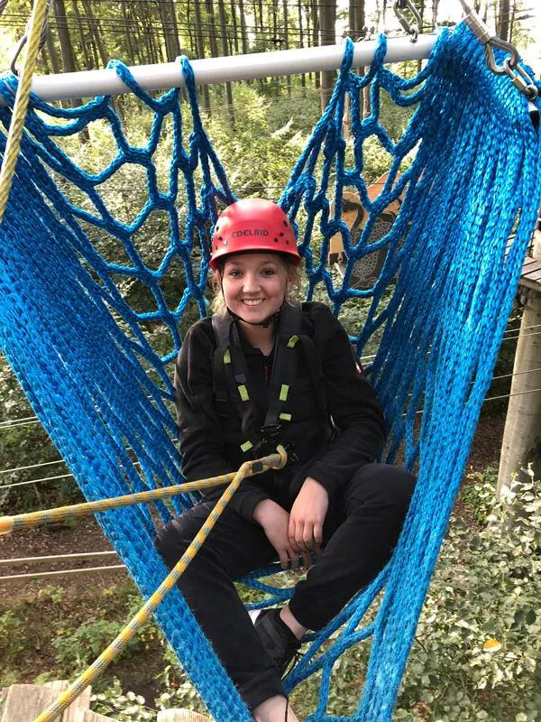 Lächendes Kind in einem blauen Netz auf einem Baumkletterpark, trägt einen roten Helm und Sicherheitsgeschirr.