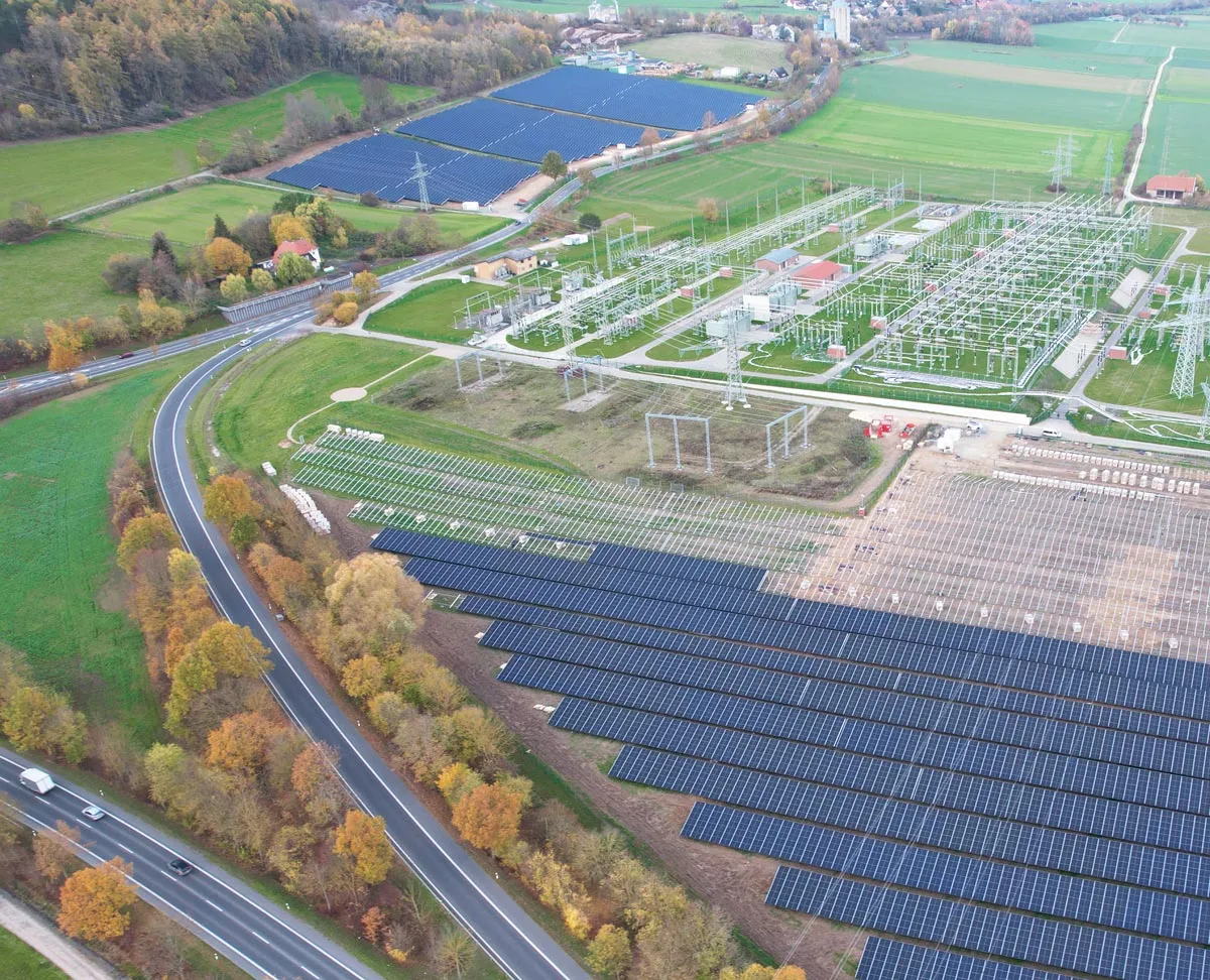 Luftaufnahme eines Gebiets mit Solarzellen, Stromleitungen und grünen Flächen, umgeben von Bäumen und Straßen.