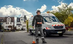 Mann in Arbeitskleidung nutzt ein Messgerät auf der Straße, im Hintergrund ein Einsatzfahrzeug und Wohnhäuser.