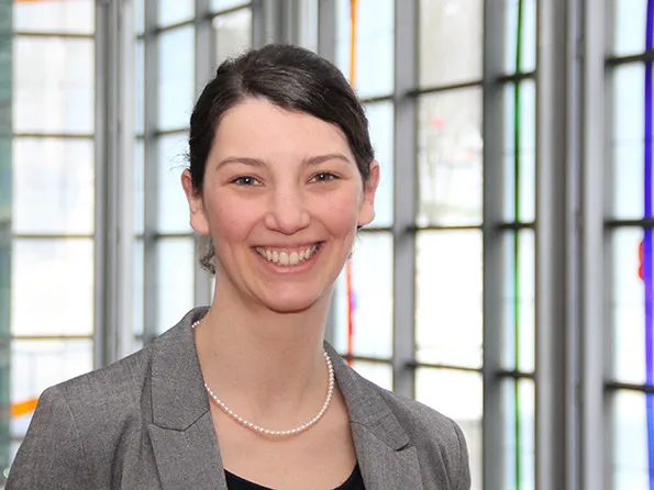 Frau mit lächelndem Gesicht, trägt einen grauen Blazer und eine Perlenkette, Hintergrund aus farbigem Glas.