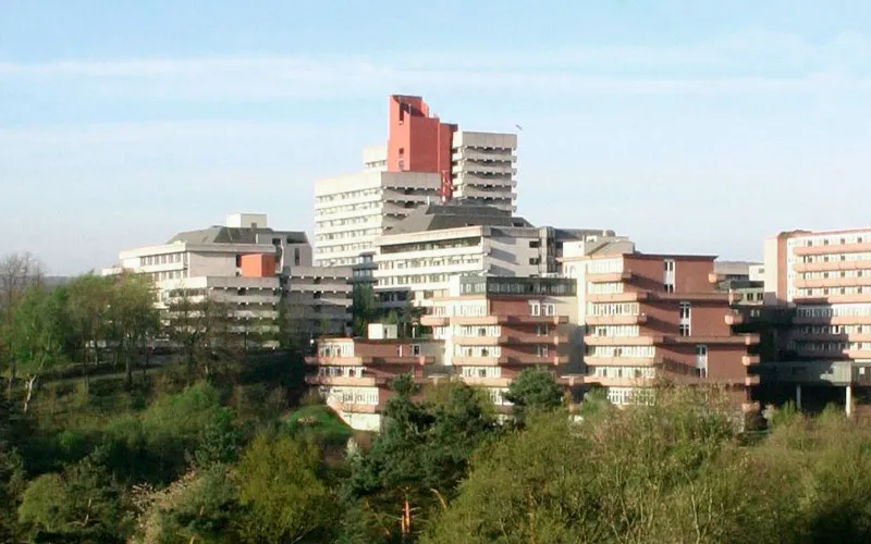 Stadtansicht mit modernen Gebäuden und begrünter Umgebung, Himmel im Hintergrund.