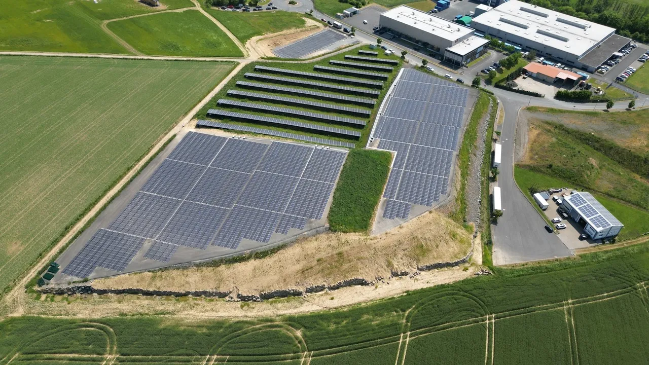  Luftaufnahme einer großen Solarpark-Anlage mit vielen Solarmodulreihen, umgeben von grünen Feldern und einem Industriegebiet.