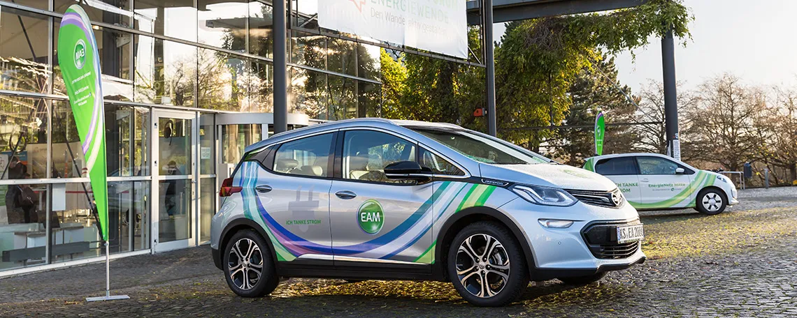 Elektroauto mit EAM-Logo vor einem modernen Gebäude, begleitende Fahnen im Hintergrund.