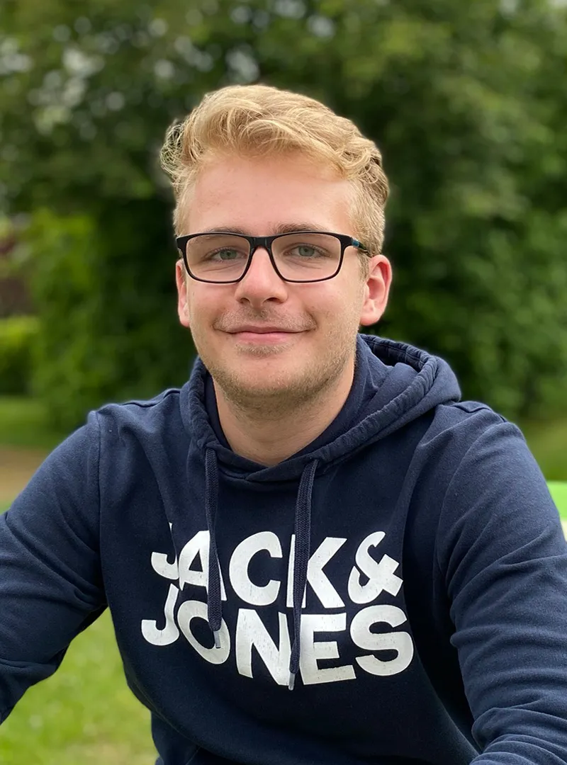 Junger Mann mit Brille, lächelnd, trägt einen dunkelblauen Hoodie mit Schriftzug, steht in einer grünen Umgebung.