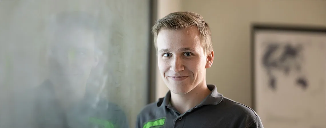 Ein junger Mann mit kurzem, blondem Haar lächelt vor einer transparenten Oberfläche, die einen schemenhaften Hintergrund zeigt.