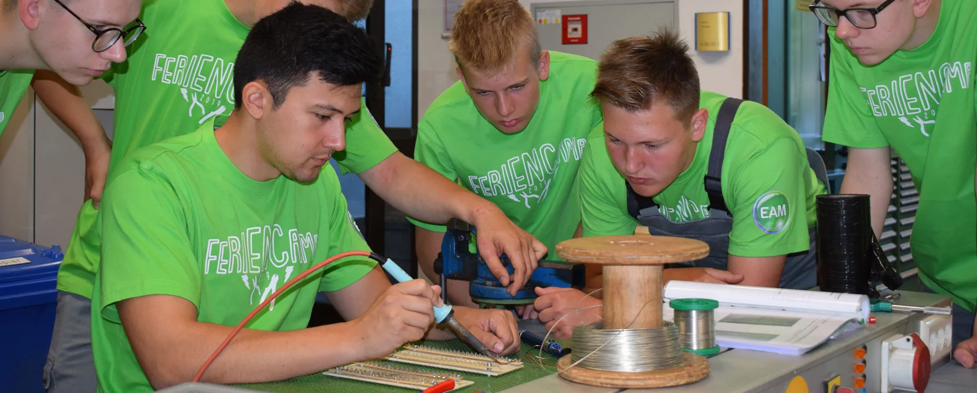 Sechs Jugendliche in grünen T-Shirts arbeiten gemeinsam an einem elektronischen Projekt mit Werkzeugen und Materialien.