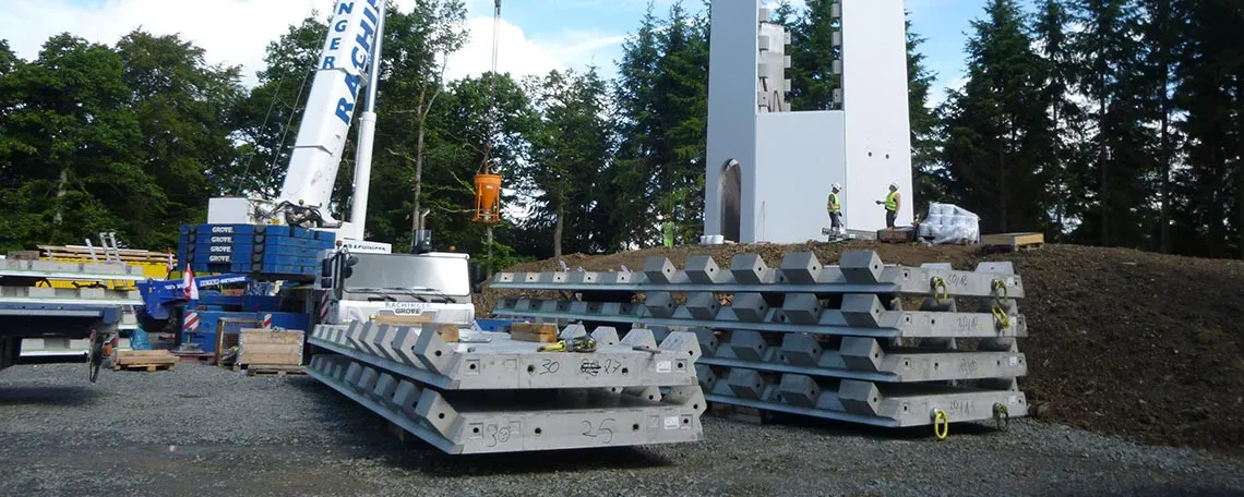 Bauplatz mit Betonblöcken, Kran und Arbeiter, umgeben von Bäumen.