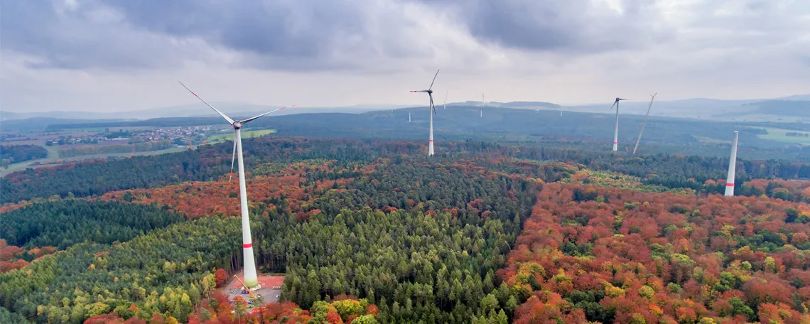 Luftaufnahme eines herbstlichen Waldes mit Windkraftanlagen, umgeben von buntem Laub und sanften Hügeln im Hintergrund.