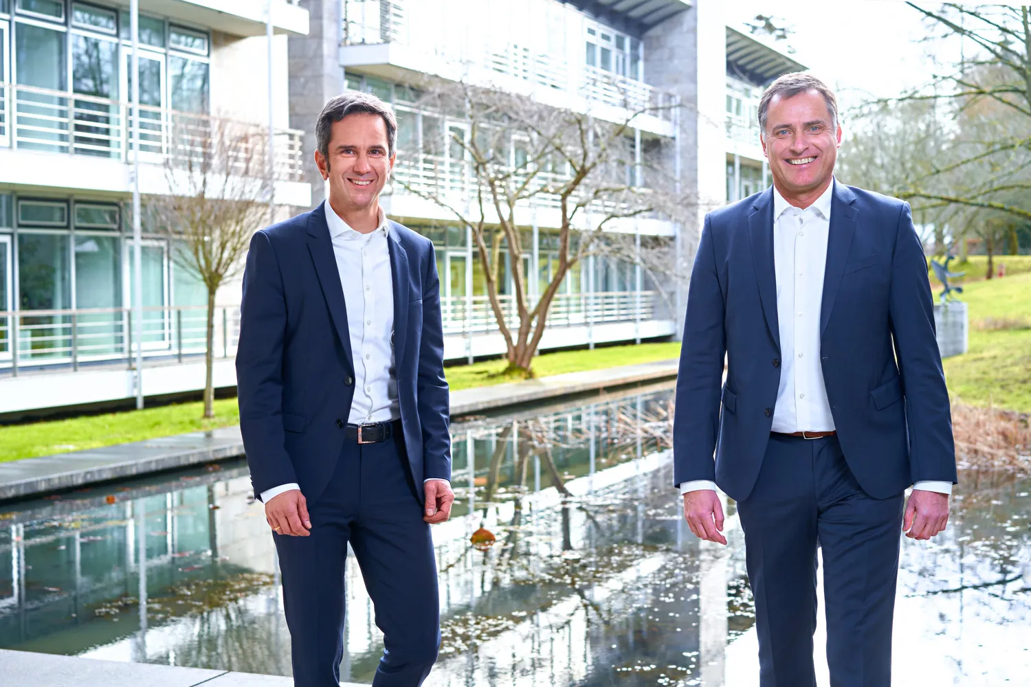 Zwei Geschäftsleute in Anzügen stehen lächelnd vor einem modernen Gebäude am Wasser.