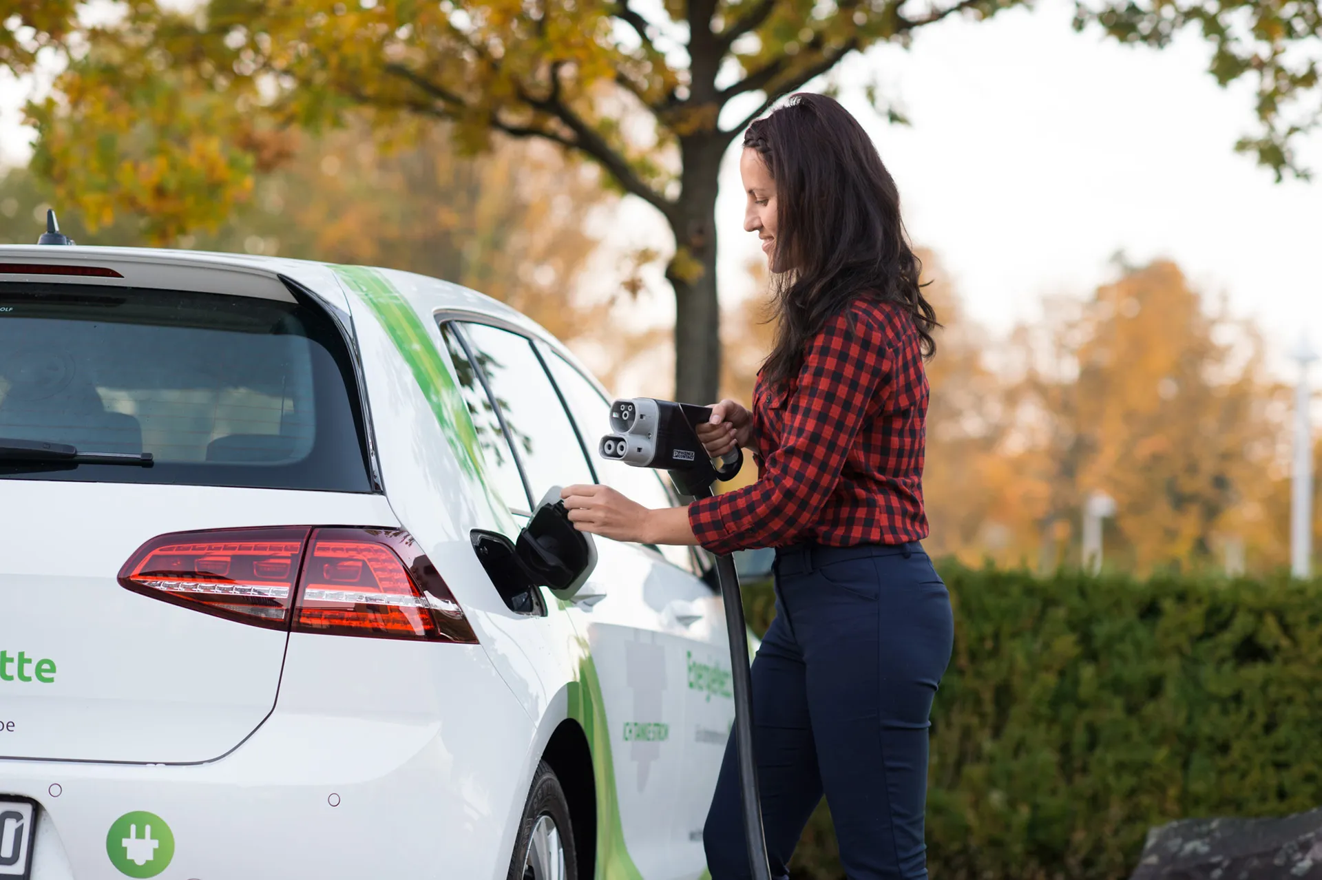  Eine lächelnde Frau in rot-kariertem Hemd und Jeans steckt ein Ladekabel in den Ladeanschluss eines weißen Elektroautos von Volkswagen.