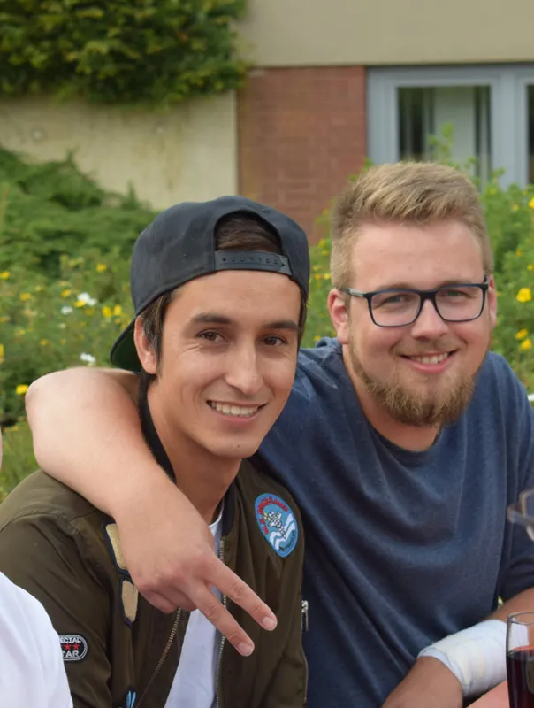 Zwei junge Männer lachen und posieren zusammen, umarmt und mit lässigen Outfits, vor einer grünen Kulisse.