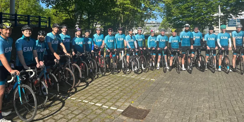 Eine Gruppe Radfahrer in blauen EAM-Trikots und Helmen stehen mit ihren Rennrädern auf einem gepflasterten Platz.