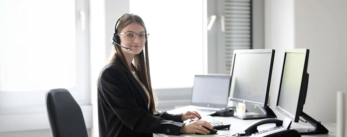 Junge Frau mit Headset sitzt an einem Schreibtisch mit mehreren Monitoren und arbeitet am Computer.