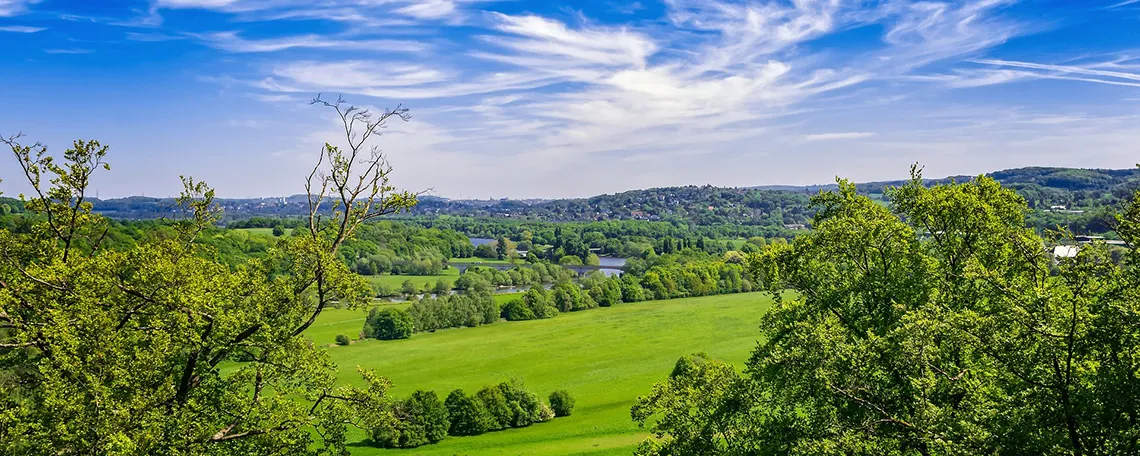 Weitläufige grüne Landschaft mit Bäumen und einem blauen Himmel, sanften Hügeln und einem Fluss im Hintergrund.