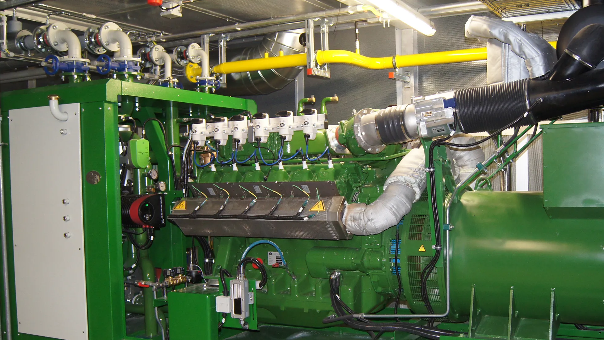 Großer grüner Generator mit Rohren, Ventilen und elektrischen Komponenten in einem technischen Raum.